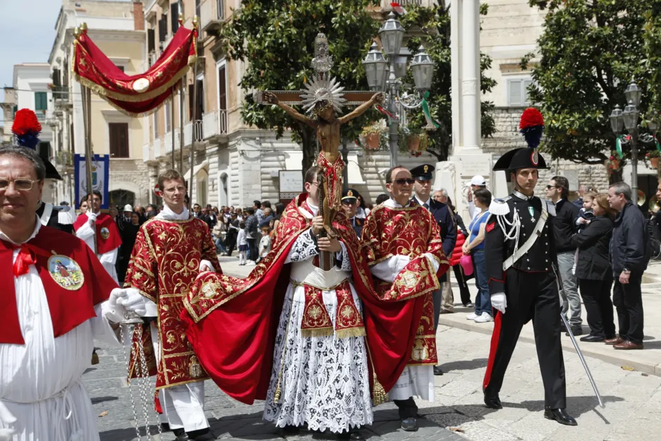 Sacerdoti in piviale rosso portano il Crocifisso in processione per strada, affiancati da un carabiniere in alta uniforme