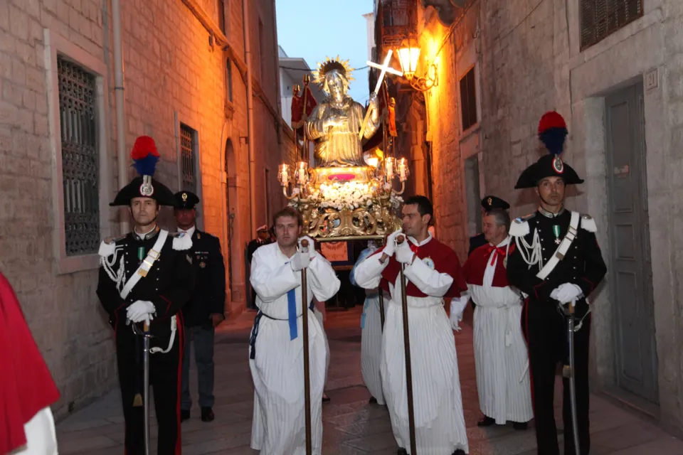 La statua illuminata di San Nicola Pellegrino avanza in un vicolo del centro storico, preceduta da confratelli e carabinieri in alta uniforme, al crepuscolo