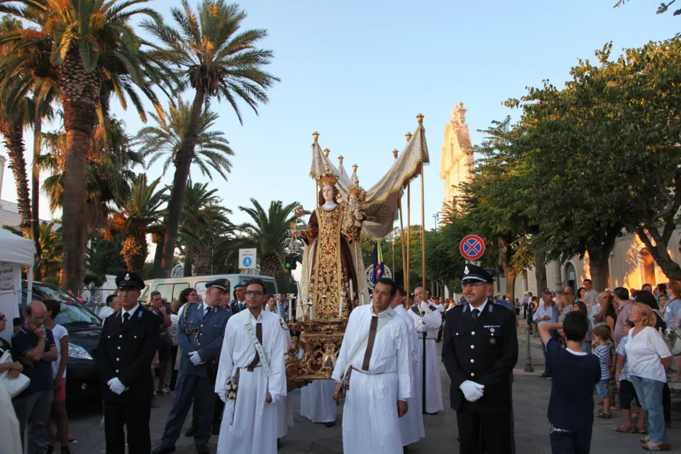 La statua della Madonna del Carmine sotto il baldacchino portata in processione in una strada alberata con palme, preceduta da confratelli e vigili in uniforme