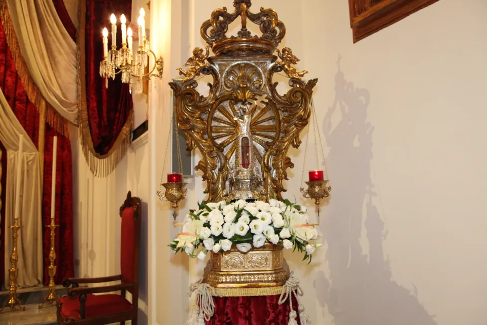 Reliquiario in argento e metallo dorato riccamente decorato, con fiori bianchi alla base, esposto accanto a drappi rossi e un candelabro