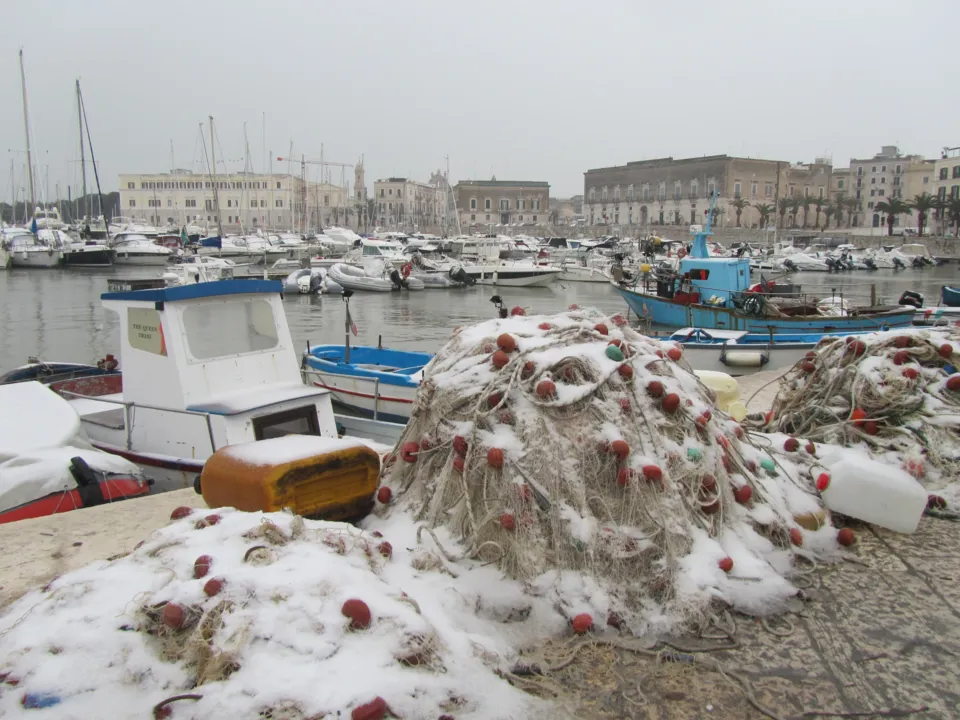 Reti da pesca coperte di neve sulla banchina, con barche da pesca e da diporto ormeggiate e i palazzi del lungoporto sullo sfondo, durante la nevicata del febbraio 2012