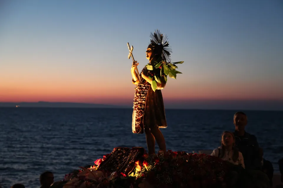 Statua lignea di San Nicola Pellegrino con croce e fronde, adorna di fiori, ripresa al tramonto con il mare e la linea di costa sullo sfondo
