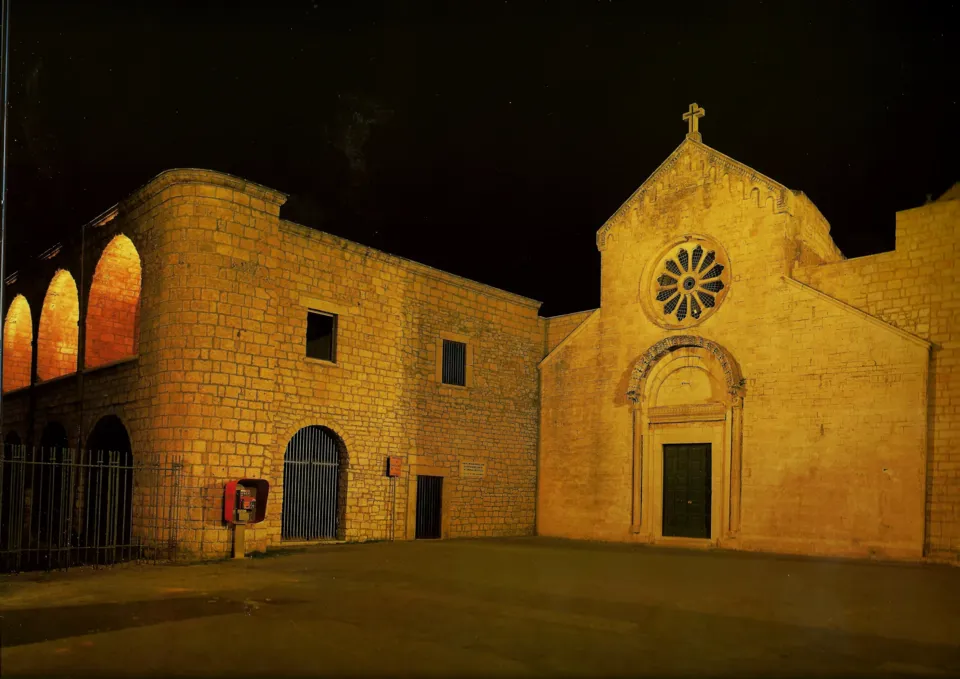 Facciata in pietra della chiesa con rosone e portale, accanto all'edificio del monastero con loggia ad archi, in illuminazione notturna