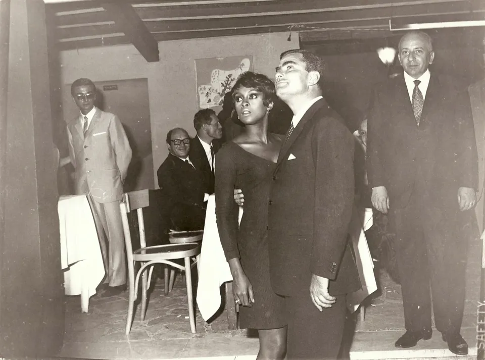 Una donna in abito scuro e un uomo in giacca posano in un locale con soffitto a travi, tra altri uomini in abito elegante e tavoli apparecchiati