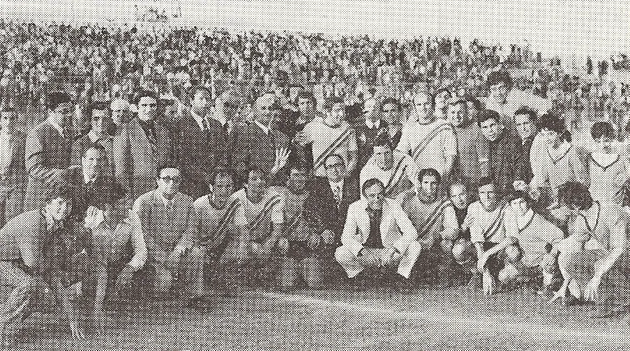 Squadra di calcio con maglie a fascia diagonale in posa con dirigenti sul campo, davanti alla tribuna gremita