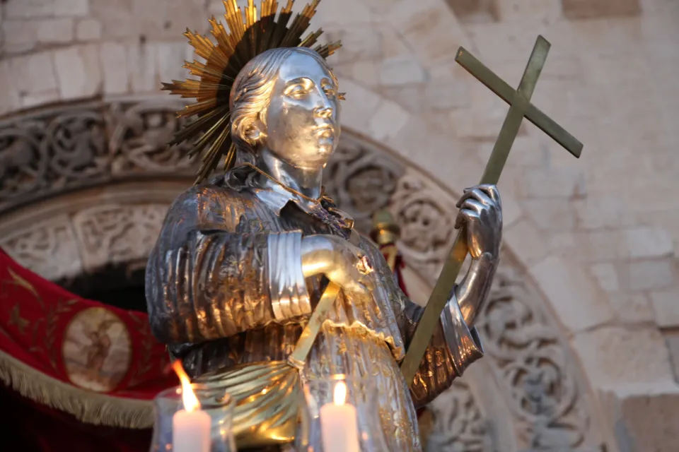 Primo piano della statua in argento di San Nicola Pellegrino con aureola e croce, con il portale scolpito della Cattedrale e candele sullo sfondo