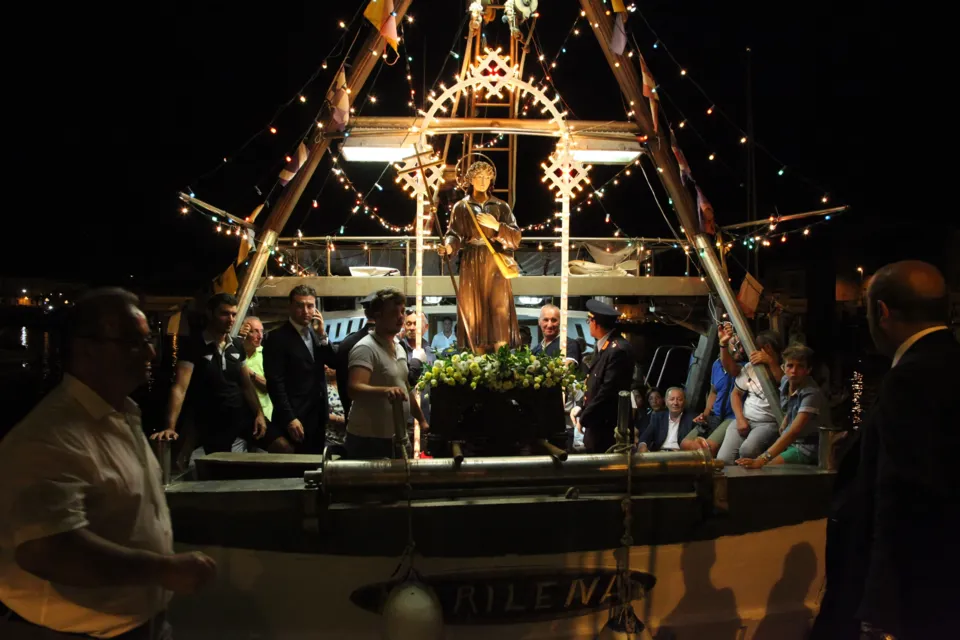 La statua di San Nicola Pellegrino a bordo di un peschereccio addobbato con luci di notte, con persone a bordo, per la processione a mare
