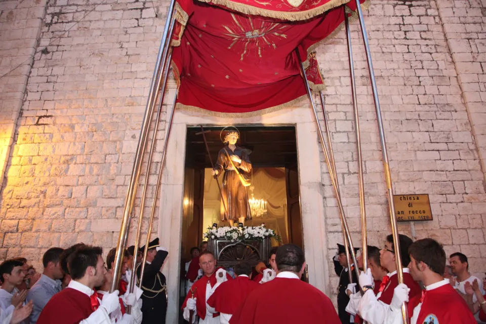 Una statua di santo con il bastone esce sotto il baldacchino rosso dal portale della chiesa di San Rocco, fra confratelli in mozzetta rossa; sul muro la targa "Chiesa di San Rocco sec