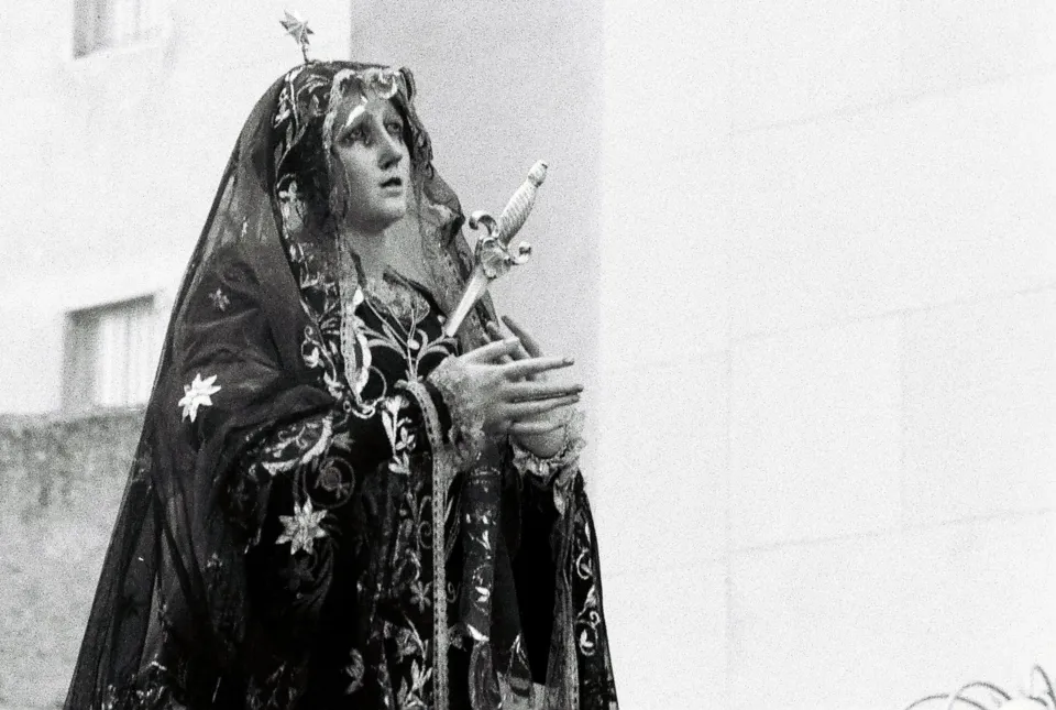 Statua della Madonna Addolorata con manto scuro ricamato, aureola e spada nel petto, portata in processione davanti a una facciata chiara