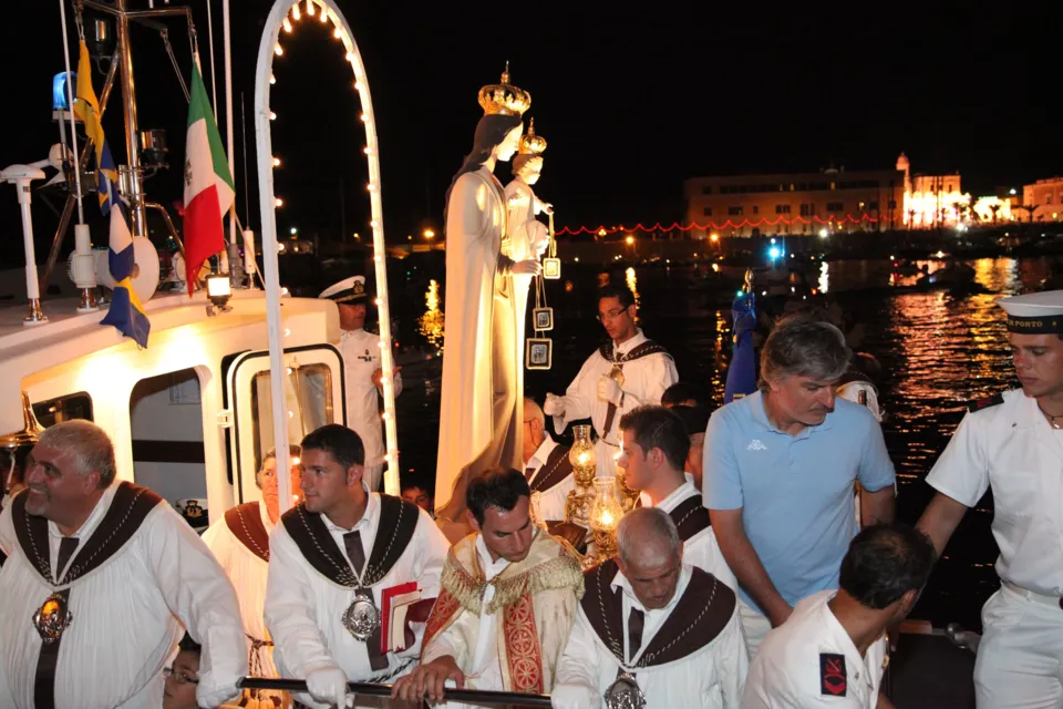 La statua della Madonna del Carmine sotto un arco luminoso a bordo dell'imbarcazione, con sacerdote, confratelli e marinai; luci della città sullo sfondo