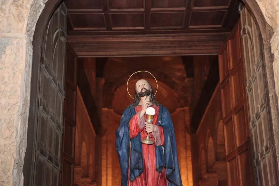 La statua di Gesù con il calice esce dal portone di una chiesa, inquadrata dagli stipiti e dal soffitto ligneo dell'interno