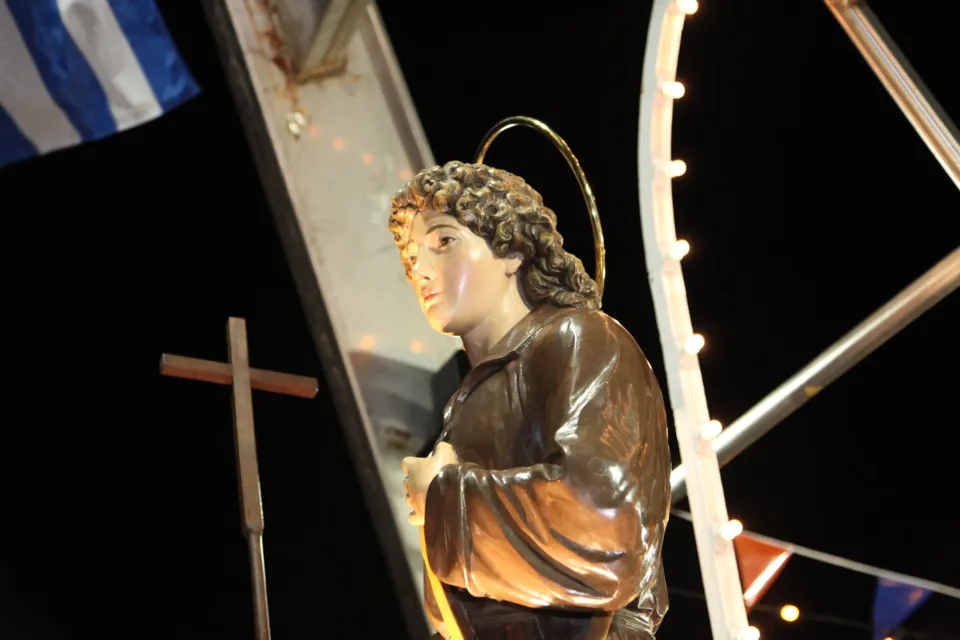 La statua di San Nicola Pellegrino, giovane con aureola e croce, a bordo dell'imbarcazione sotto un arco di luci, con la bandiera greca