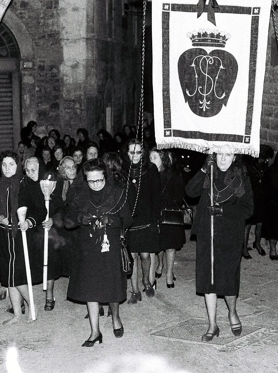 Stendardo con monogramma sormontato da corona portato in processione davanti a un gruppo di donne in abiti neri con ceri