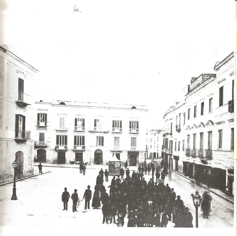 Fotografia d'epoca di una strada con palazzi ottocenteschi, lampioni e numerosi passanti in abiti scuri