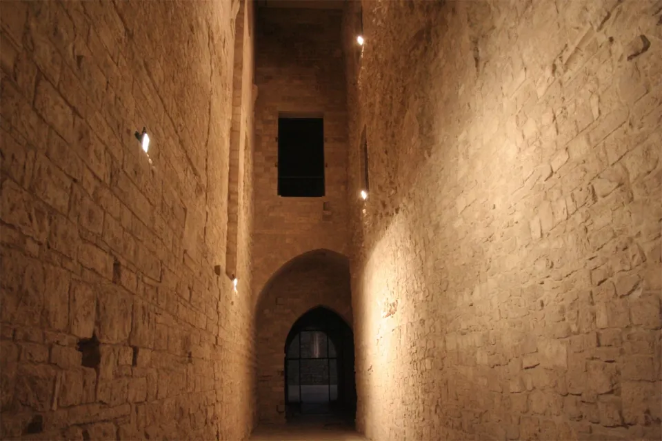 Corridoio stretto e alto tra muri in pietra del Castello Svevo, illuminato da faretti, con un portale ogivale in fondo