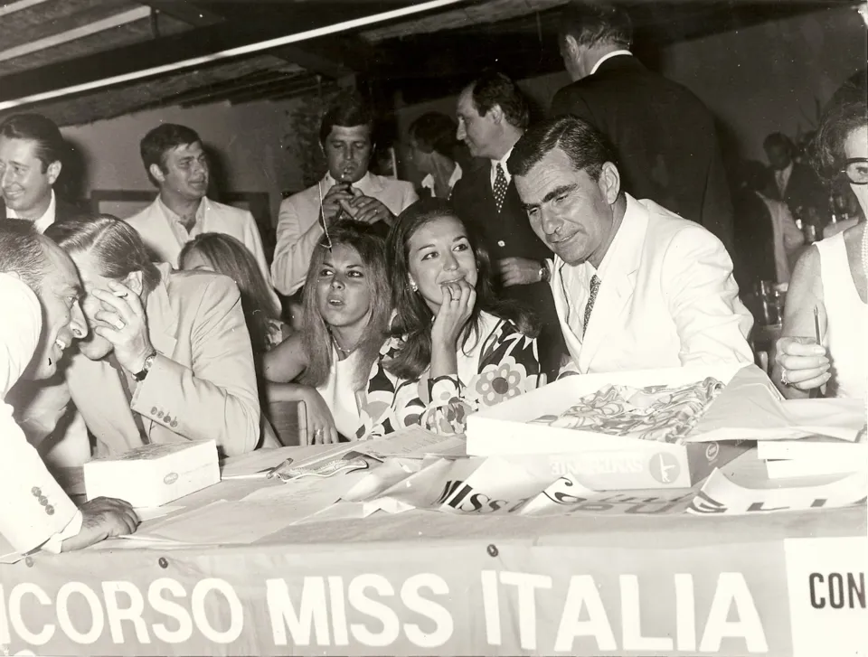 Giovani donne e uomini seduti a un tavolo con lo striscione 'Concorso Miss Italia', con altri ospiti in piedi alle spalle