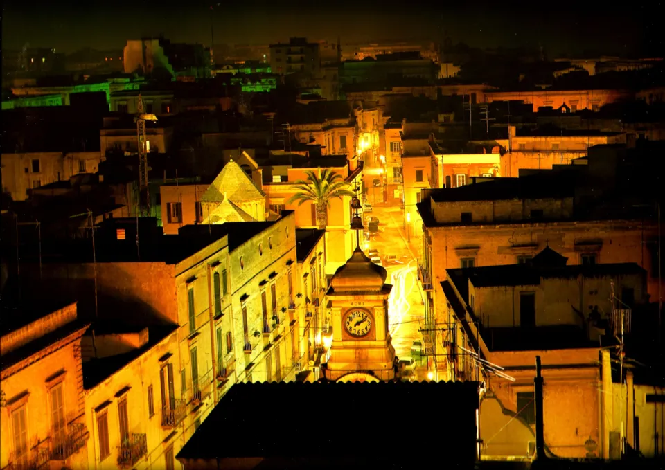 Panorama notturno dei tetti con una torre dell'orologio illuminata e una strada che prosegue verso il fondo