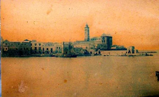 Foto d'epoca virata seppia del fronte a mare di Trani, con le case basse lungo la costa e il campanile della Cattedrale