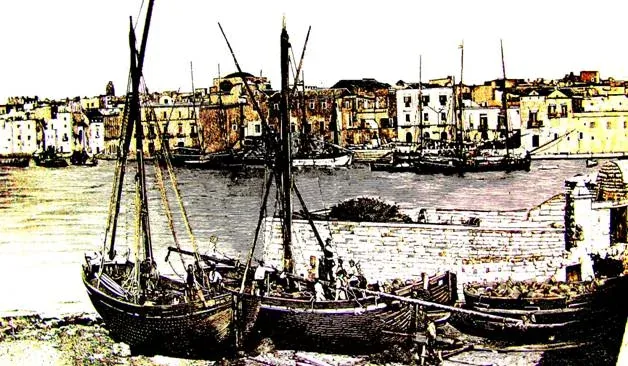 Foto d'epoca colorata a mano con velieri da pesca in secca e ormeggiati lungo la banchina, con le case del fronte porto sullo sfondo
