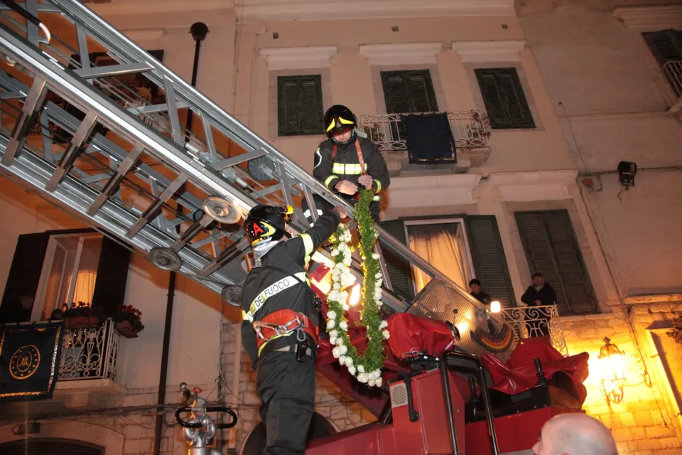 Un vigile del fuoco sale sull'autoscala portando la corona di fiori, davanti alla facciata di un palazzo