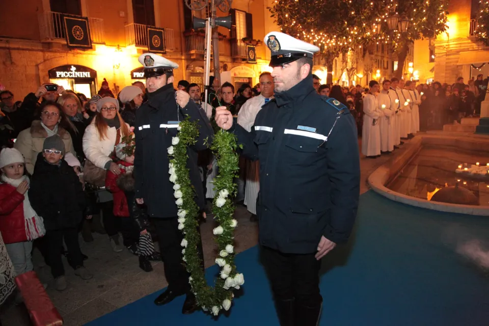 Due agenti della Polizia Municipale portano una corona di fiori bianchi tra la folla, verso il monumento all'Immacolata